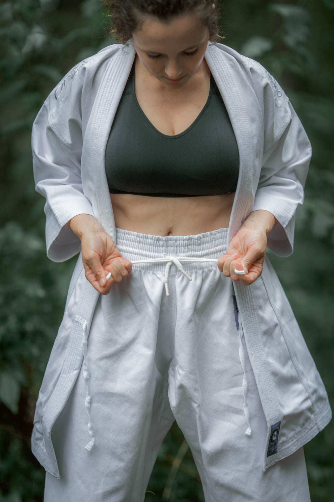Person tying white karate pants wearing a white jacket and sports bra out side in the rain.